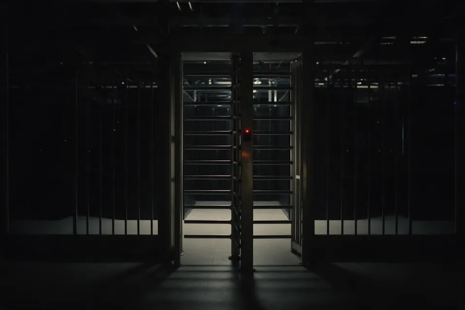 A single turnstile gate at the entrance to a dark server hall, one small red indicator lit on it.