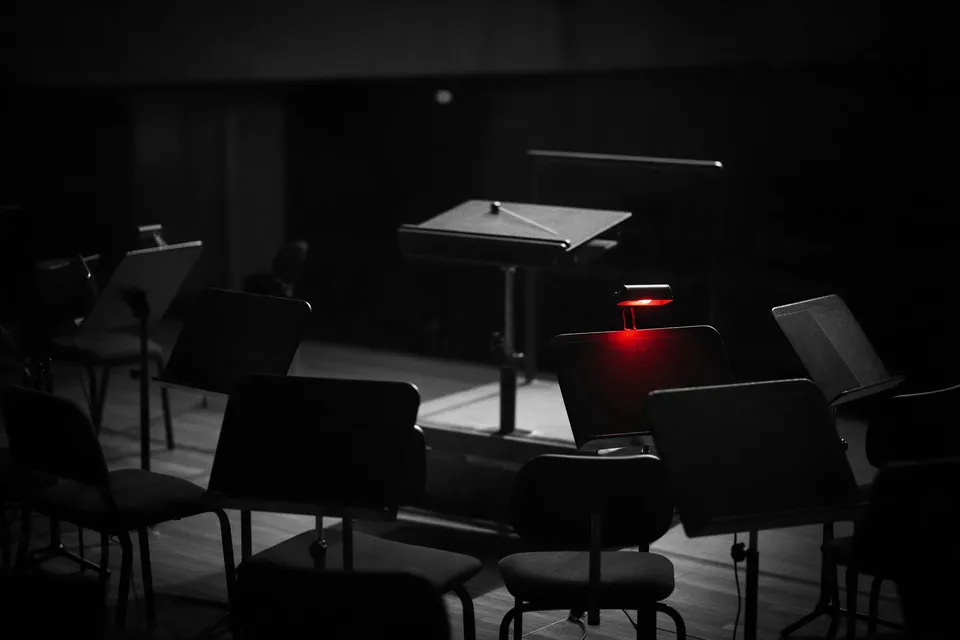 An empty conductor's podium in a dark concert hall, the players' chairs and music stands in front of it, one stand lamp glowing red.