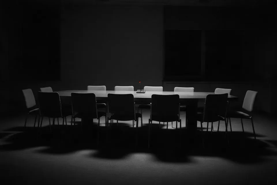 An empty conference room at night, twelve chairs around a long table, one small desk microphone with a red light.